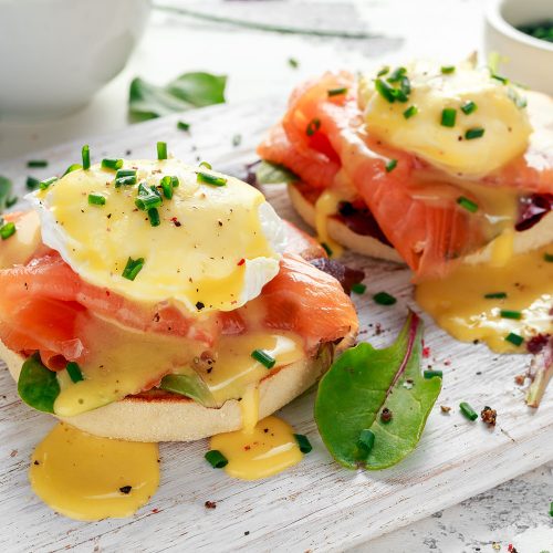 Eggs Benedict on english muffin with smoked salmon, lettuce salad mix and hollandaise sauce on white board.